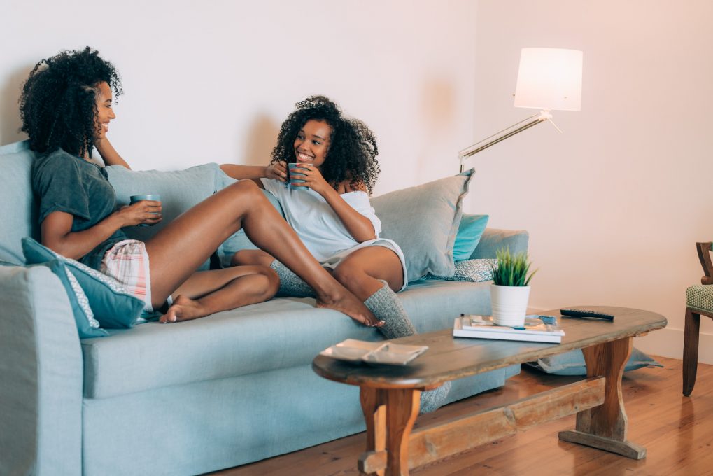 Happy young two black women sitting in the couch drinking coffee 'n